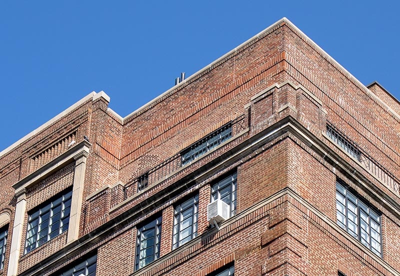 A close-up, low-angle view of the upper corner of a multi-story brick building set against a clear blue sky.