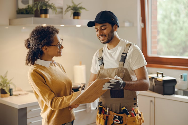 A professional service technician in overalls and a tool belt reviewing paperwork on a clipboard with a homeowner in her kitchen.